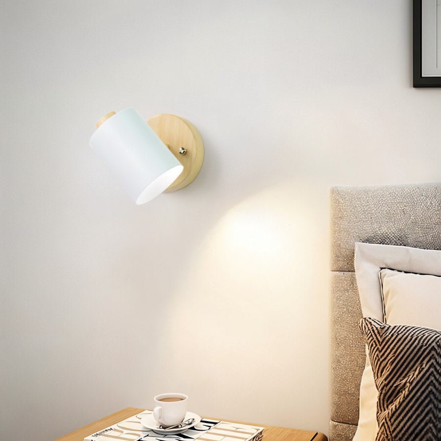 Clérachic wall lamp with a white metal shade on a wooden backplate casting a warm pool of light beside a bed and nightstand.