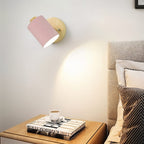 Clérachic wall lamp with a pink metal shade and wooden backplate throwing a soft spotlight over a bedside table and headboard.