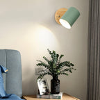 Clérachic wall lamp with a green metal shade on a round wooden backplate lighting a bedside table with a vase of eucalyptus.