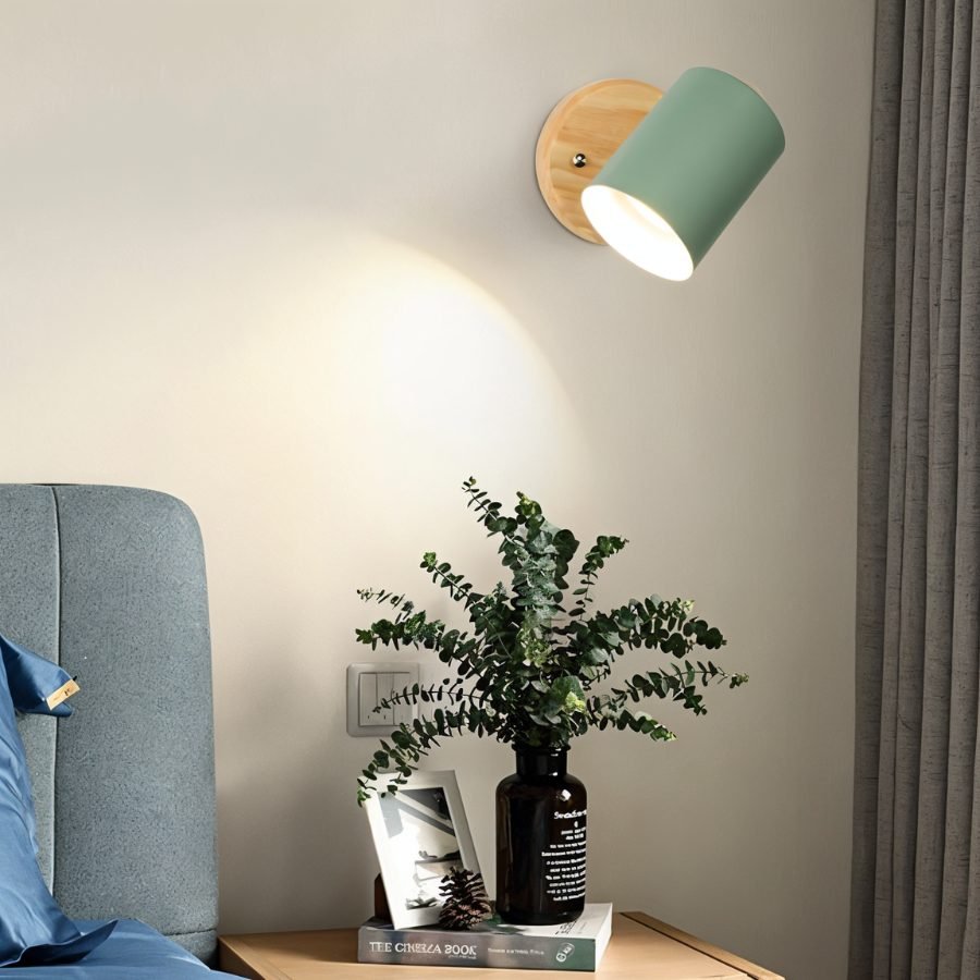 Clérachic wall lamp with a green metal shade on a round wooden backplate lighting a bedside table with a vase of eucalyptus.