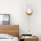 Clérachic wall lamp with a white metal shade, wooden backplate and a gold pull chain, mounted above a wooden bedside table.