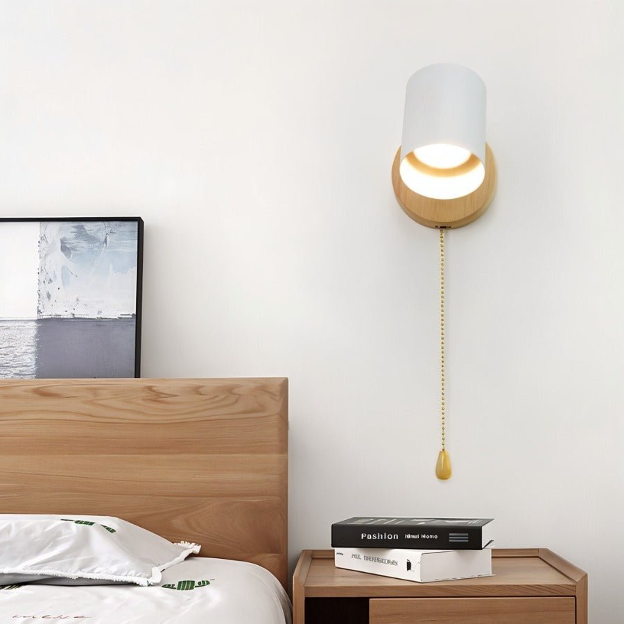 Clérachic wall lamp with a white metal shade, wooden backplate and a gold pull chain, mounted above a wooden bedside table.