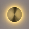 Lit brass disk wall light from Clérachic, a warm halo of light glowing around its edge on a pale grey wall.
