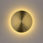 Lit brass disk wall light from Clérachic, a warm halo of light glowing around its edge on a pale grey wall.