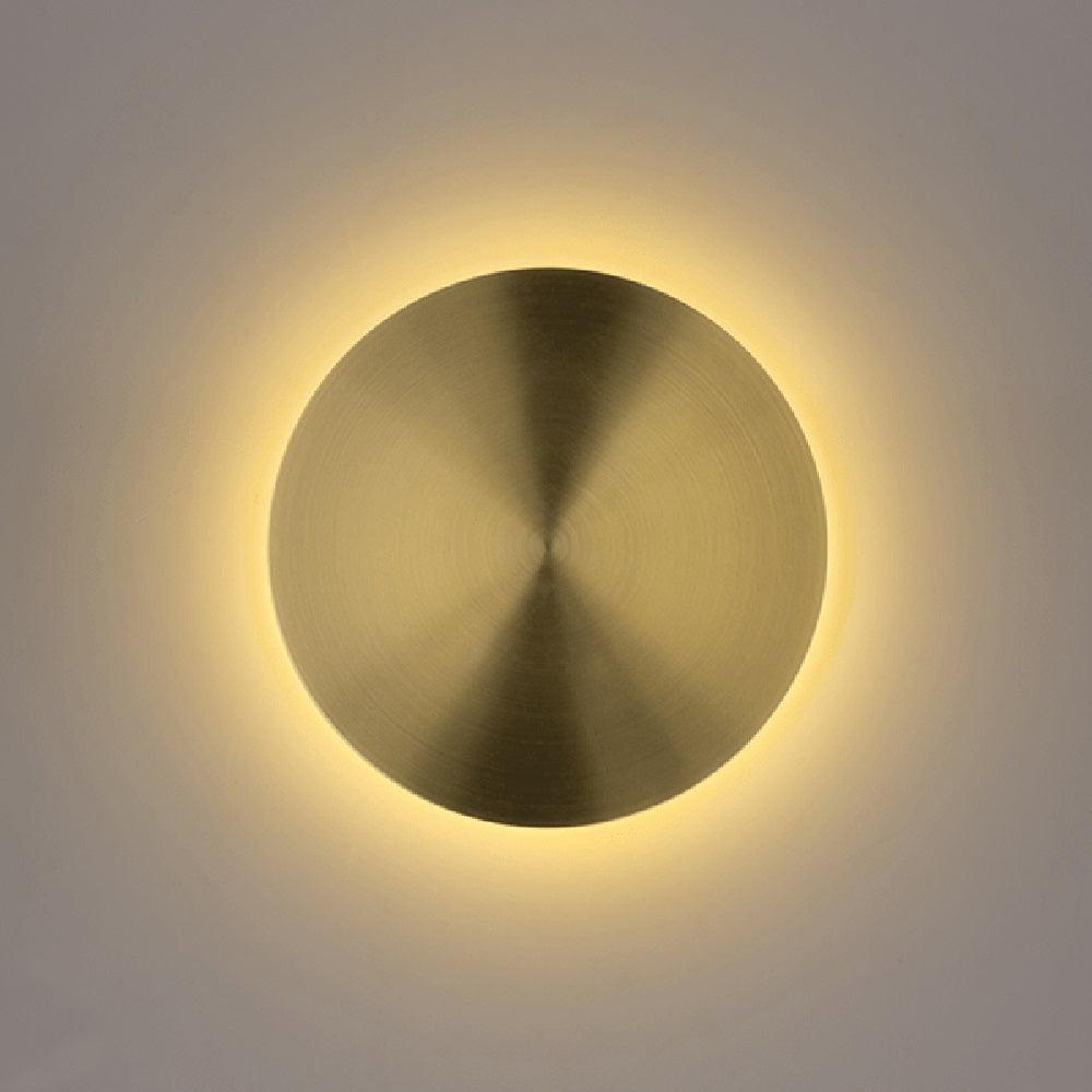 Lit brass disk wall light from Clérachic, a warm halo of light glowing around its edge on a pale grey wall.
