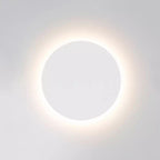 White round Clérachic wall light switched on, a bright ring of light spreading around the disk onto a pale wall.