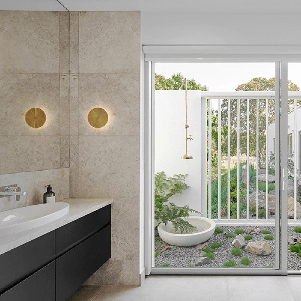 Two small brass disk wall lights from Clérachic glow above a bathroom vanity, beside a glass door to a courtyard garden.