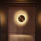 Small Clérachic disk light in brass, glowing with a halo against a dark fabric-covered wall above a padded seat.