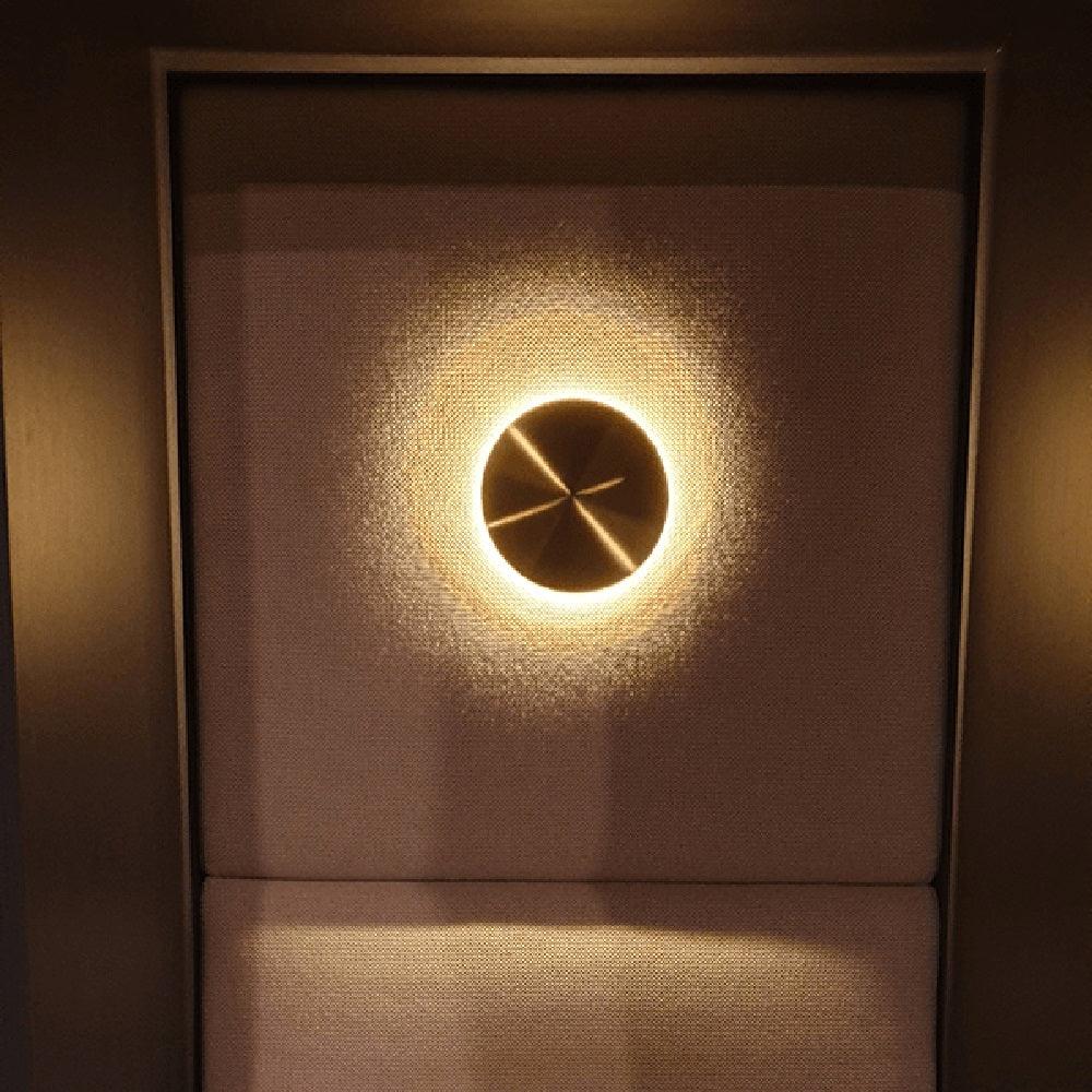 Small Clérachic disk light in brass, glowing with a halo against a dark fabric-covered wall above a padded seat.