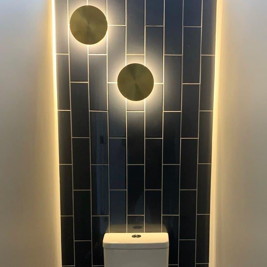 Two brass Clérachic disk lights lit on a wall of dark blue vertical tiles, with a glowing strip of light down each side.
