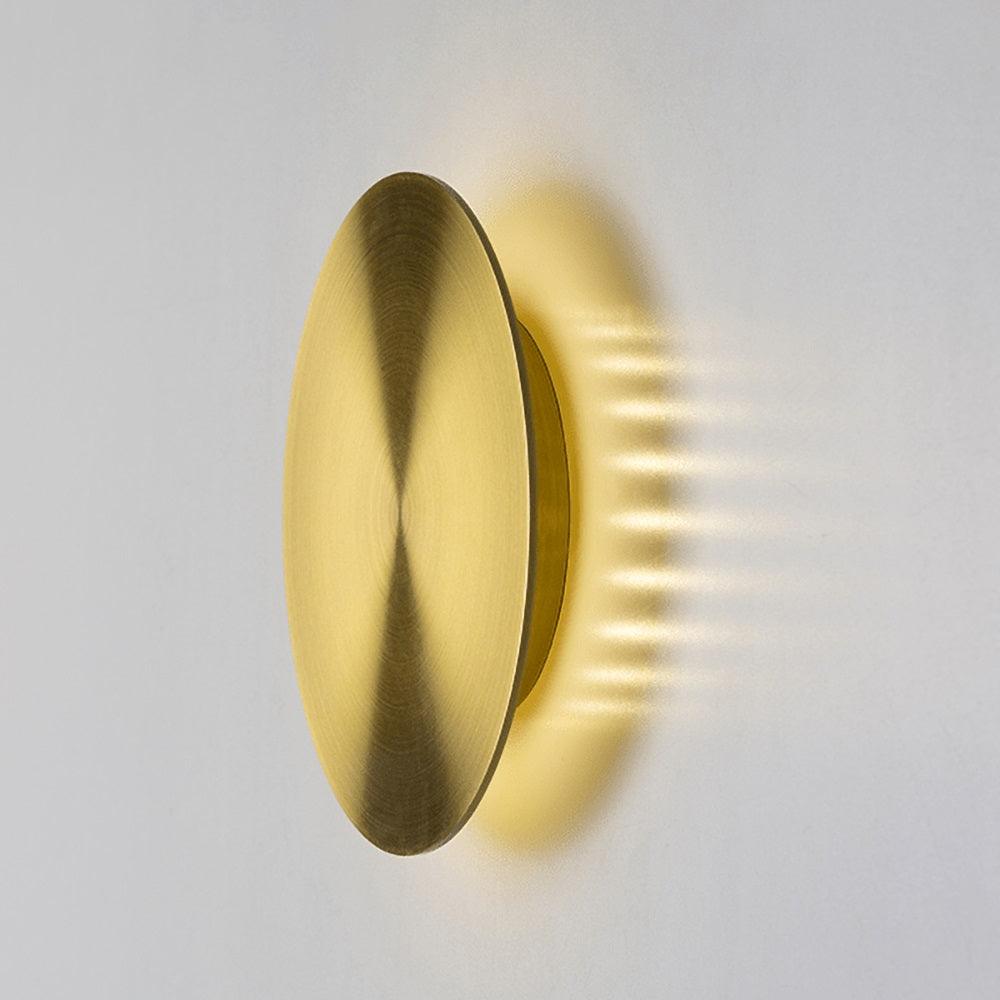 Angled view of a brushed brass Clérachic disk light on a white wall, the light spilling out sideways from behind the disk.