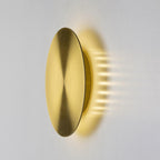 Angled view of a brushed brass Clérachic disk light on a white wall, the light spilling out sideways from behind the disk.