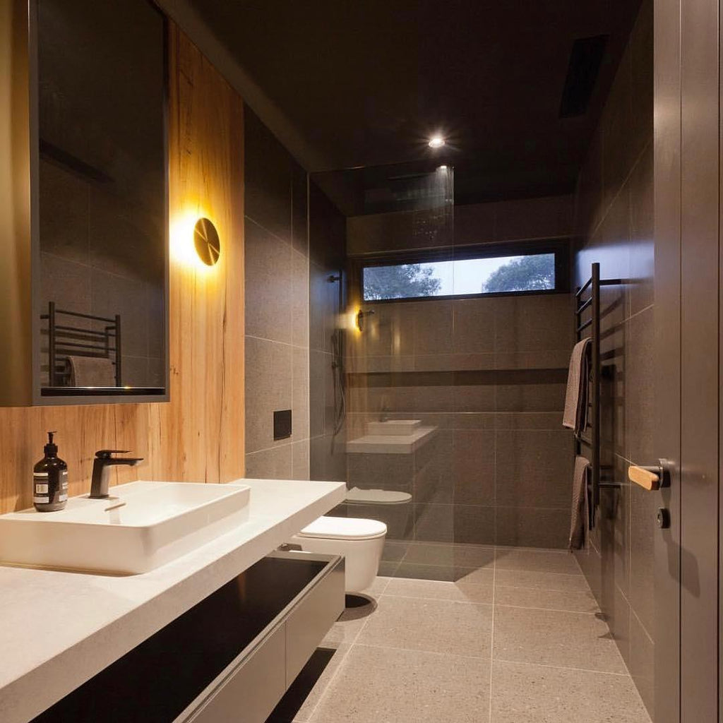 A brass Clérachic disk light glows on a wood-clad wall in a dark bathroom, beside a mirror, basin and glass shower.