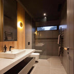 A brass Clérachic disk light glows on a wood-clad wall in a dark bathroom, beside a mirror, basin and glass shower.