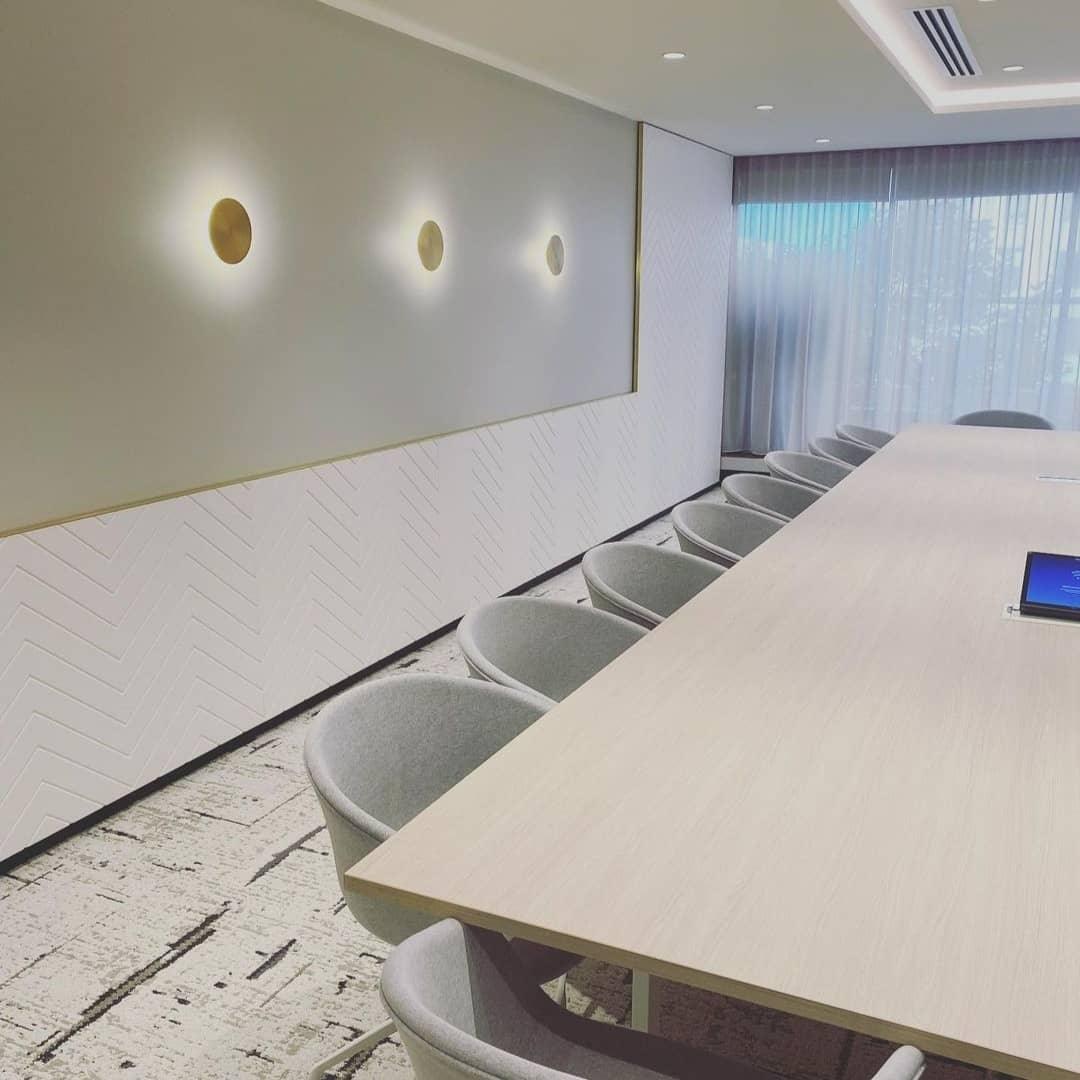 Three brass Clérachic disk lights in a row on a long meeting-room wall, above a pale table and grey chairs.