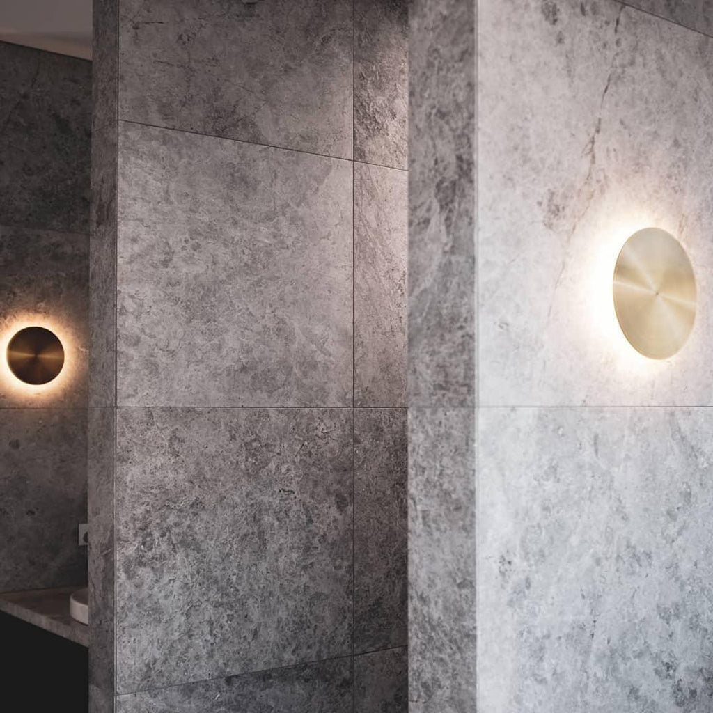 Brass Clérachic disk lights glow on a pale grey stone-effect wall, one at the left edge and one larger at the right.