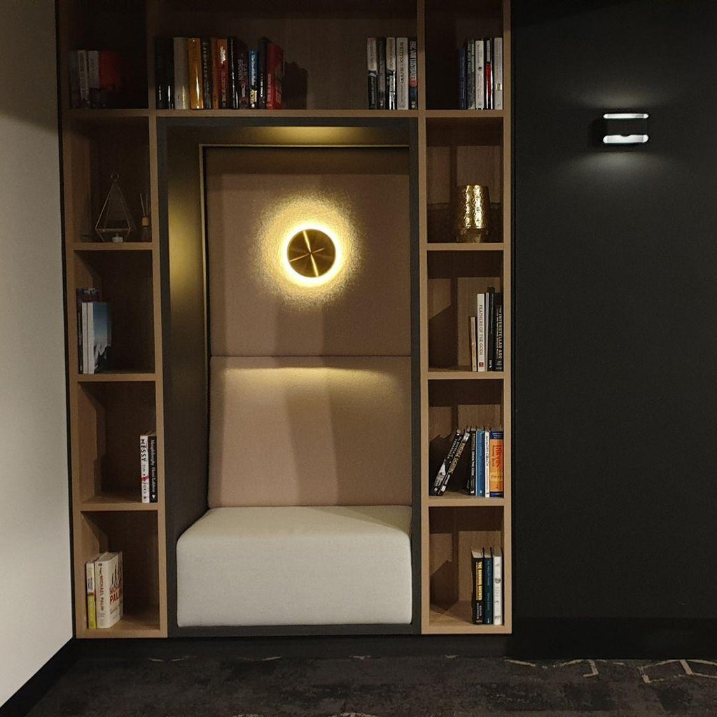 Clérachic brass disk light with a soft ring of glow, set inside a wooden bookshelf alcove with a white padded bench.