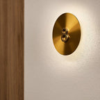 Brass disk on a textured white wall from Clérachic, with a small dark twin-ring fitting at its centre and a soft glow behind.