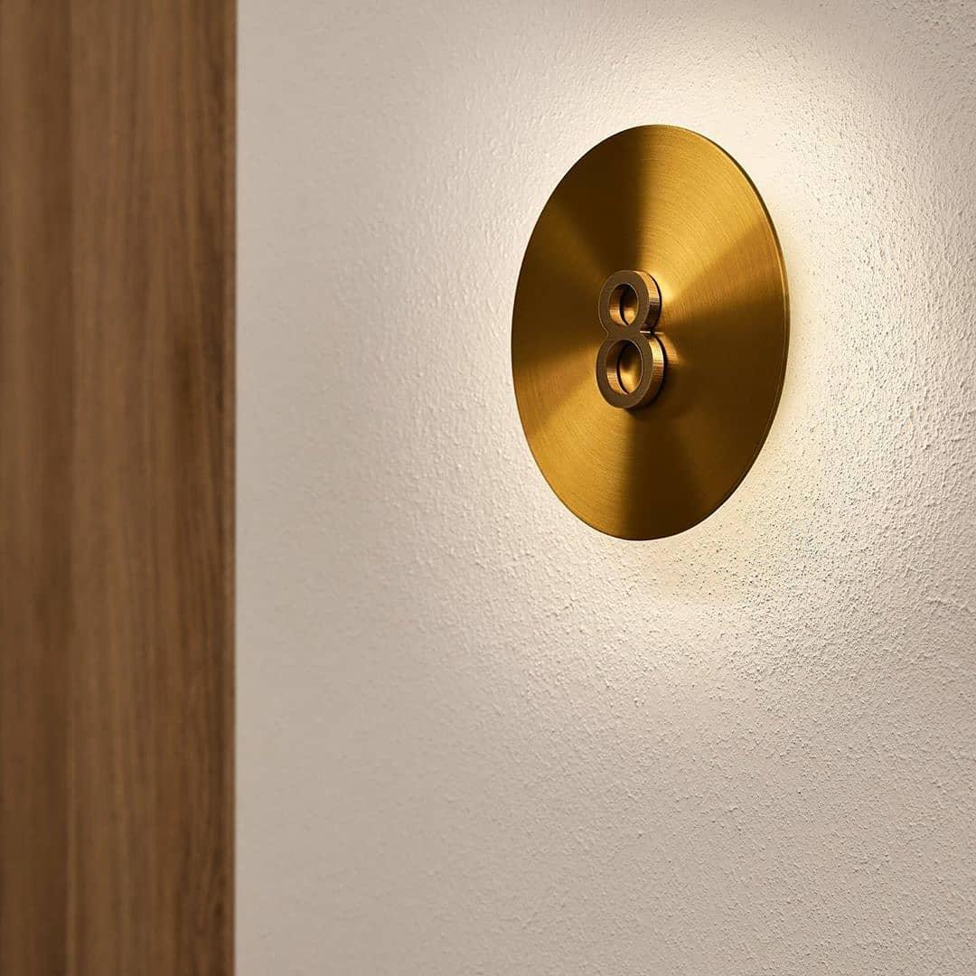 Brass disk on a textured white wall from Clérachic, with a small dark twin-ring fitting at its centre and a soft glow behind.
