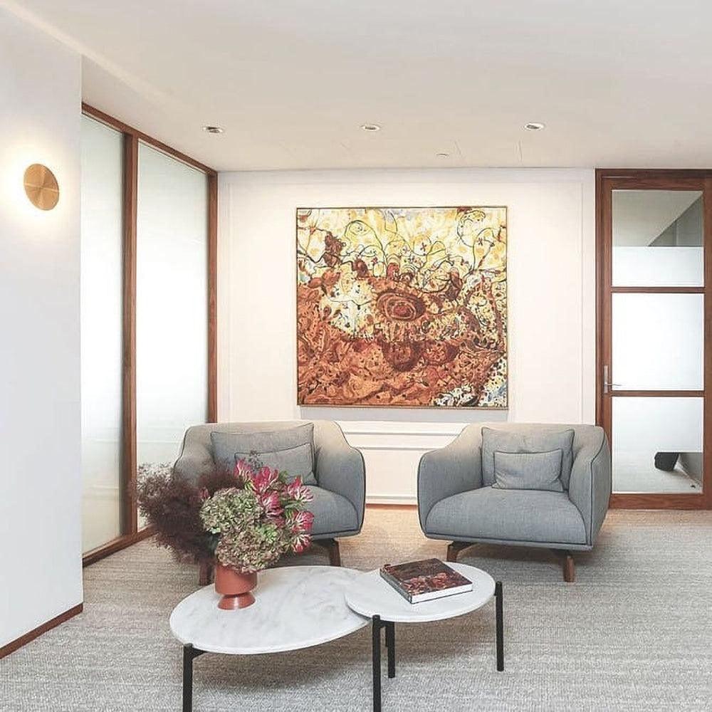Brass Clérachic disk light on a white wall of a sitting room, with two grey armchairs and round marble tables below.