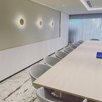 Three brass Clérachic disk lights lit along a meeting-room wall, with a long pale table and sheer curtains at the window.