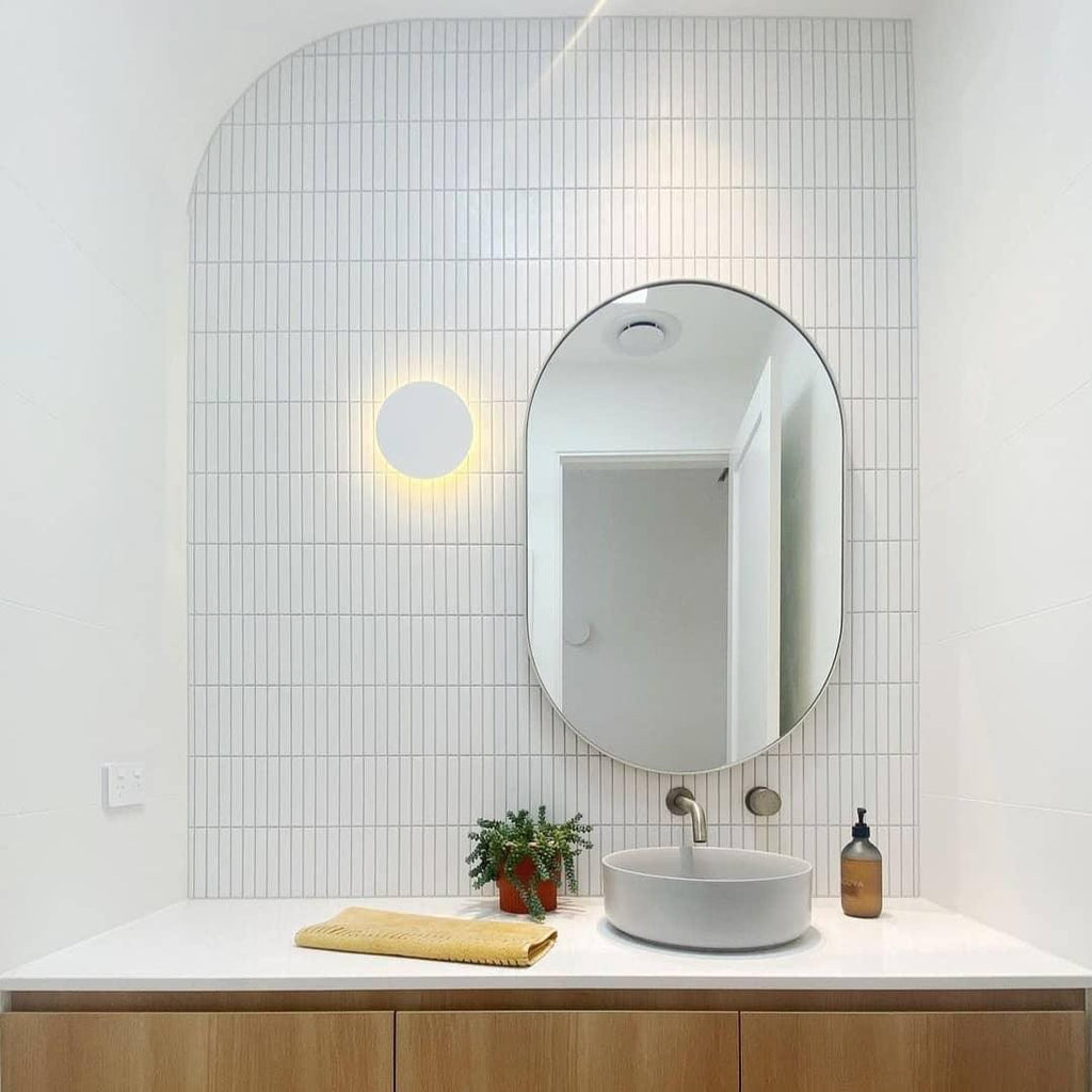 A white round Clérachic wall light glows beside an oval mirror above a white basin on a wooden vanity.
