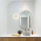 A white round Clérachic wall light glows beside an oval mirror above a white basin on a wooden vanity.