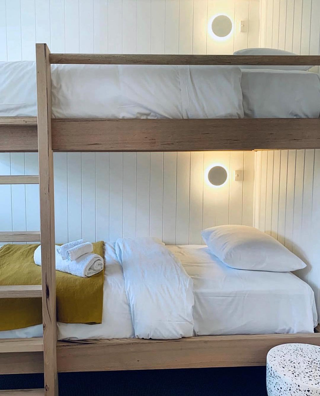 White round Clérachic wall lights glow above the upper and lower bunks of a wooden bunk bed with white bedding.
