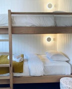 White round Clérachic wall lights glow above the upper and lower bunks of a wooden bunk bed with white bedding.