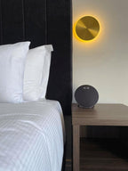 A brass Clérachic disk light with an amber glow beside a black padded headboard, above a bedside table with a round speaker.