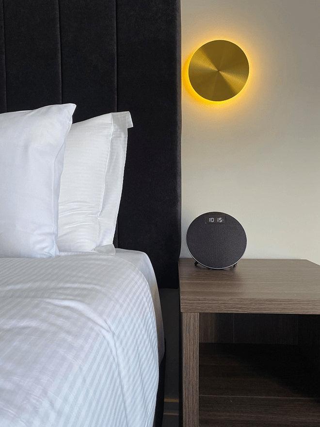 A brass Clérachic disk light with an amber glow beside a black padded headboard, above a bedside table with a round speaker.