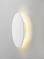 Angled view of a white Clérachic disk wall light on a pale wall, warm light spilling out behind its edge.