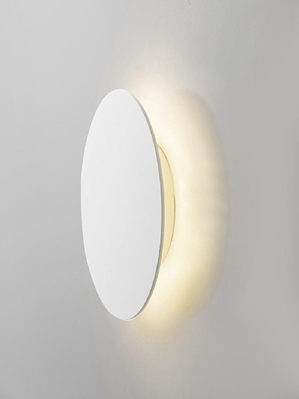 Angled view of a white Clérachic disk wall light on a pale wall, warm light spilling out behind its edge.