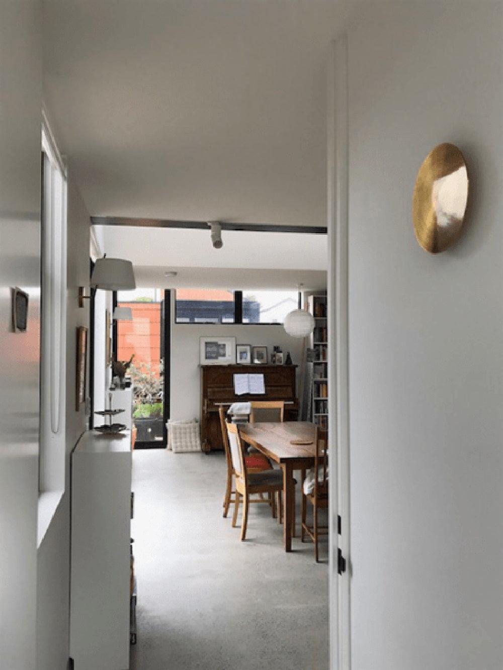 Small brass Clérachic disk light seen angled on a white wall at the end of a hallway, with a dining table and piano beyond.