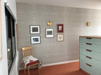 Two small brass Clérachic disk lights on a grey patterned wall above framed pictures, a wooden chair and a pale green chest.