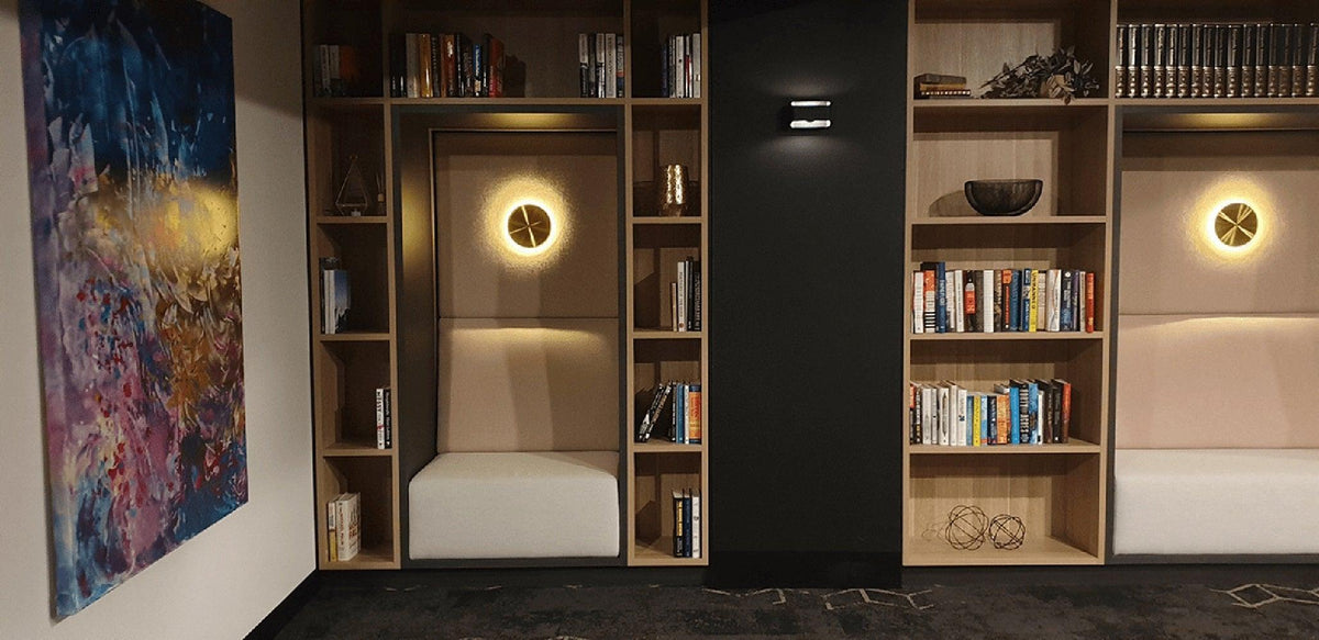 Two brass Clérachic disk lights glow in matching oak bookshelf alcoves with white seats, either side of a dark column.