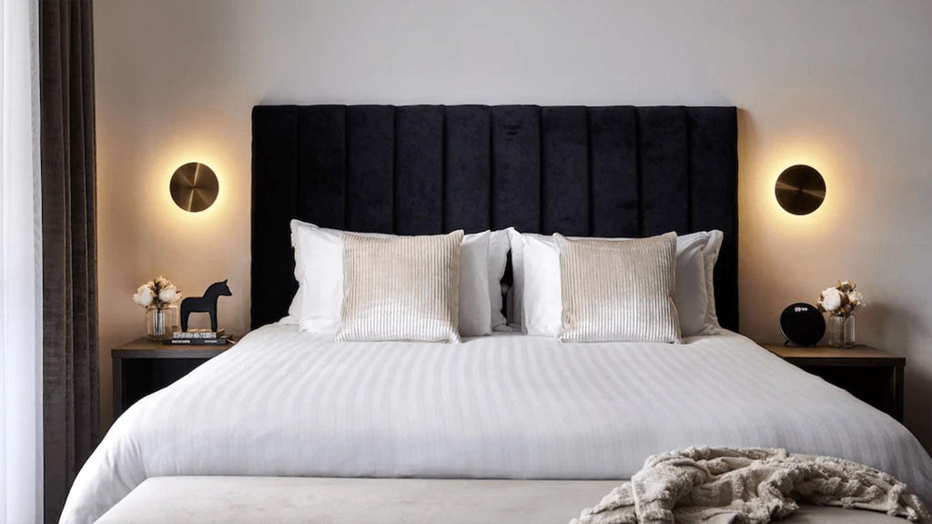 A pair of brass Clérachic disk lights flank a bed with a dark vertical-ribbed headboard and white striped bedding.