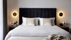 A pair of brass Clérachic disk lights flank a bed with a dark vertical-ribbed headboard and white striped bedding.