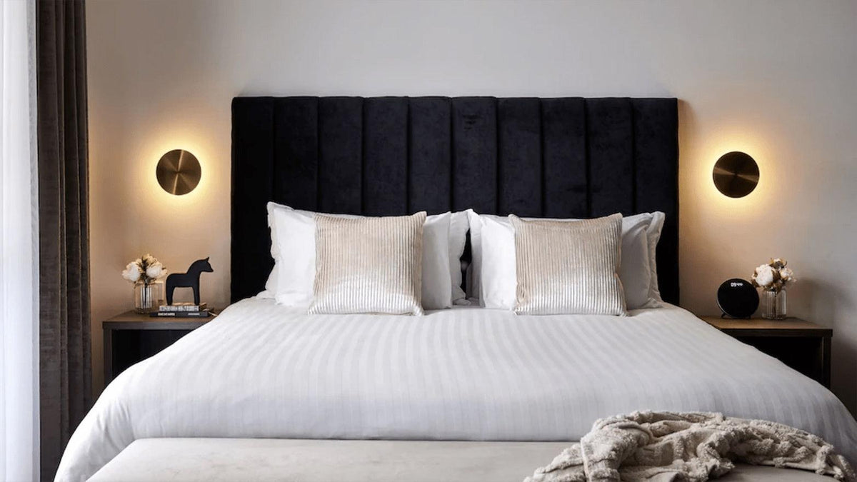 A pair of brass Clérachic disk lights flank a bed with a dark vertical-ribbed headboard and white striped bedding.
