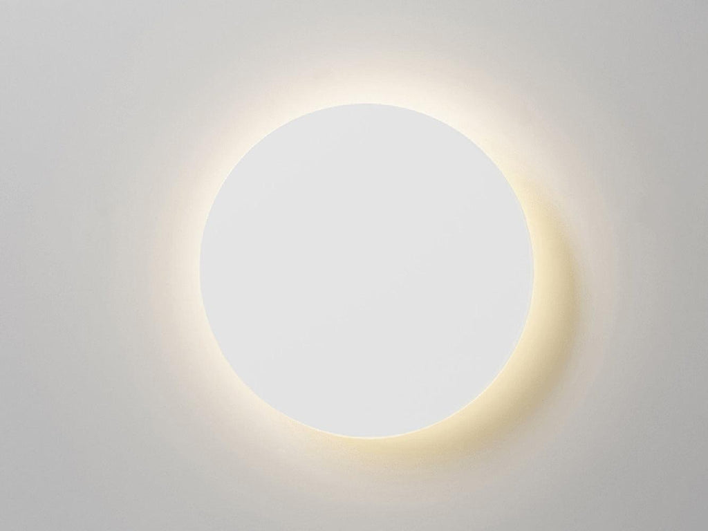 Front view of a white round Clérachic wall light lit, with a soft halo of warm light on a pale grey wall.
