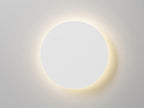 Front view of a white round Clérachic wall light lit, with a soft halo of warm light on a pale grey wall.