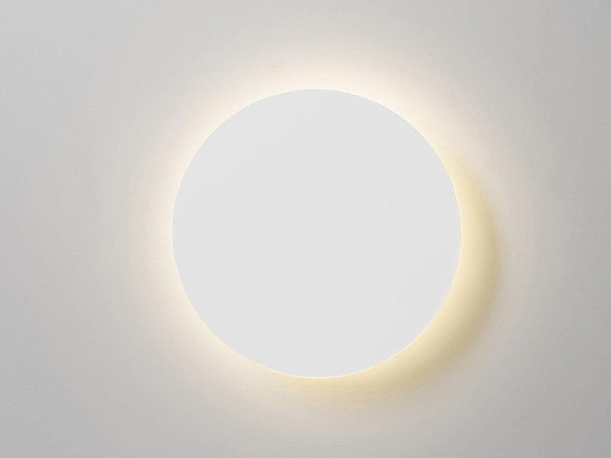 Front view of a white round Clérachic wall light lit, with a soft halo of warm light on a pale grey wall.