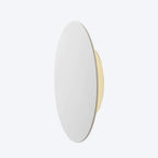 Side view of a white Clérachic disk wall light on a pale backdrop, with a brass back plate showing behind it.