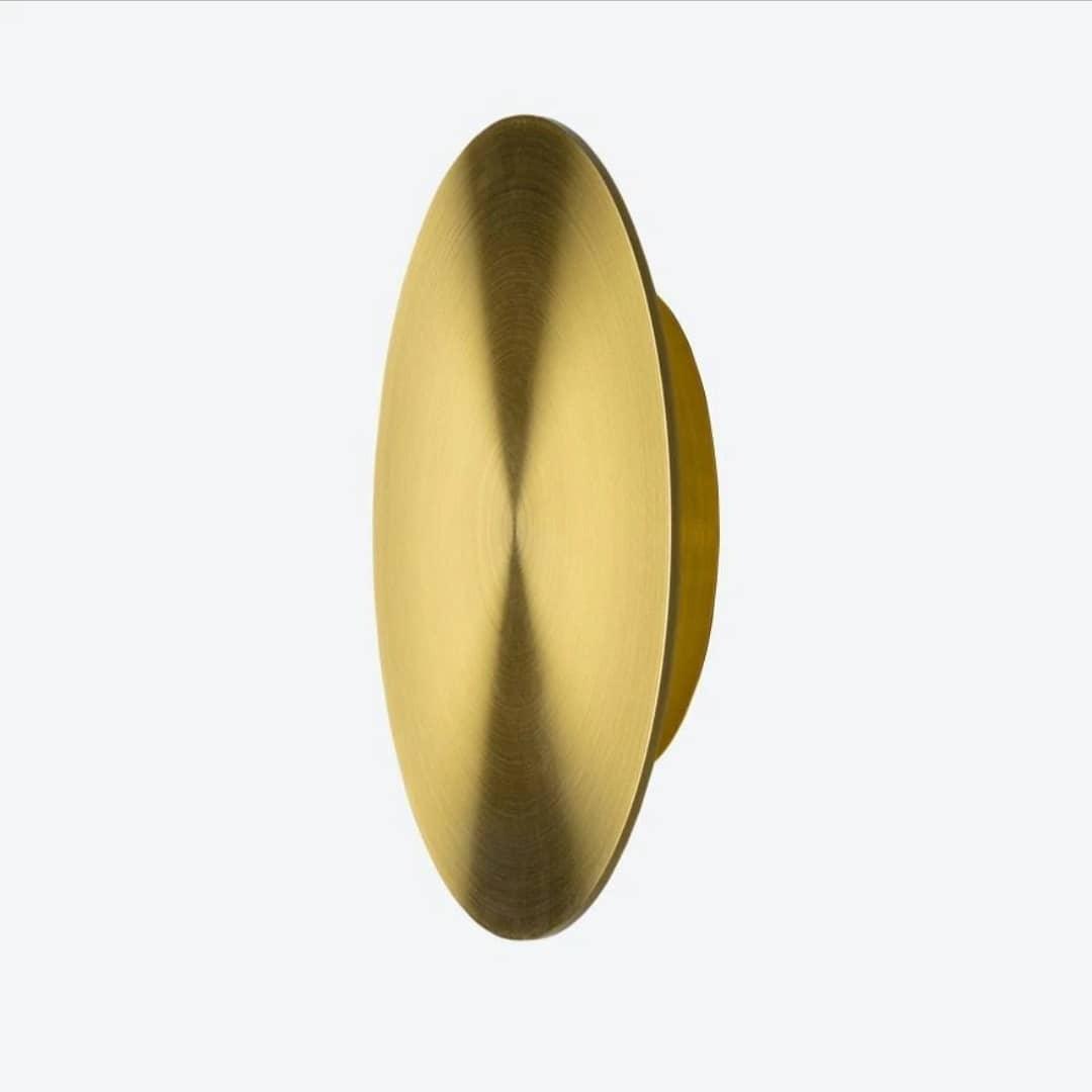 Three-quarter view of a brushed brass Clérachic disk light on a pale backdrop, with the stepped back plate visible.