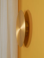 Brass Clérachic disk light seen from the side on a mustard-yellow wall, beside a cream curtain.
