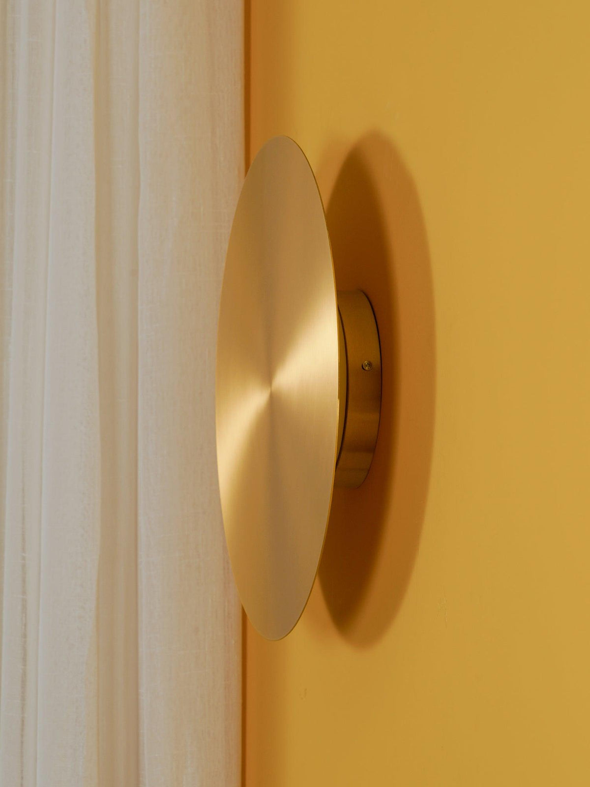 Brass Clérachic disk light seen from the side on a mustard-yellow wall, beside a cream curtain.