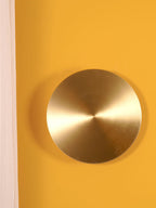 Front view of a brushed brass Clérachic disk light on a mustard-yellow wall, with a cream curtain at the left edge.