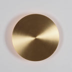 Close front view of a brushed brass Clérachic disk light, a soft pinkish glow showing around its edge on a pale wall.