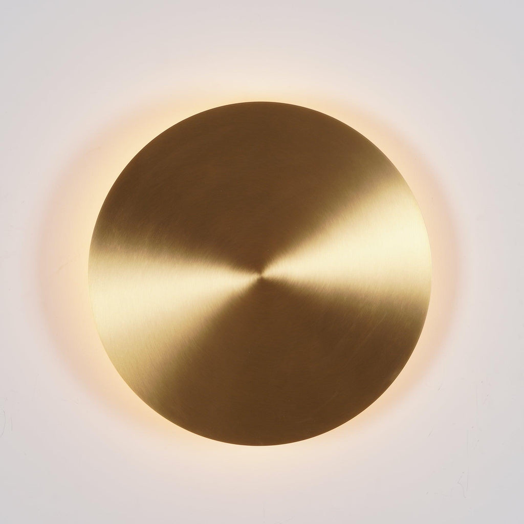 Lit brass Clérachic disk light with a warm halo of light behind its rim, on a light grey wall.