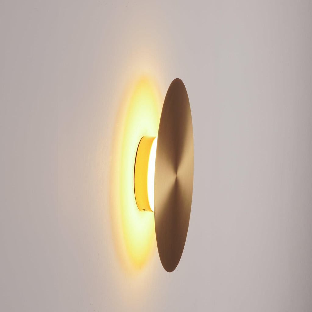 Side view of a brass Clérachic disk light lit, the amber glow shining between the disk and the pale wall behind it.